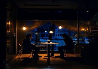 深夜咖啡馆角落两人对坐低声交谈，暖黄灯光与窗外蓝黑夜色形成强烈对比，呈现夜无疆情绪疗愈空间的温暖氛围